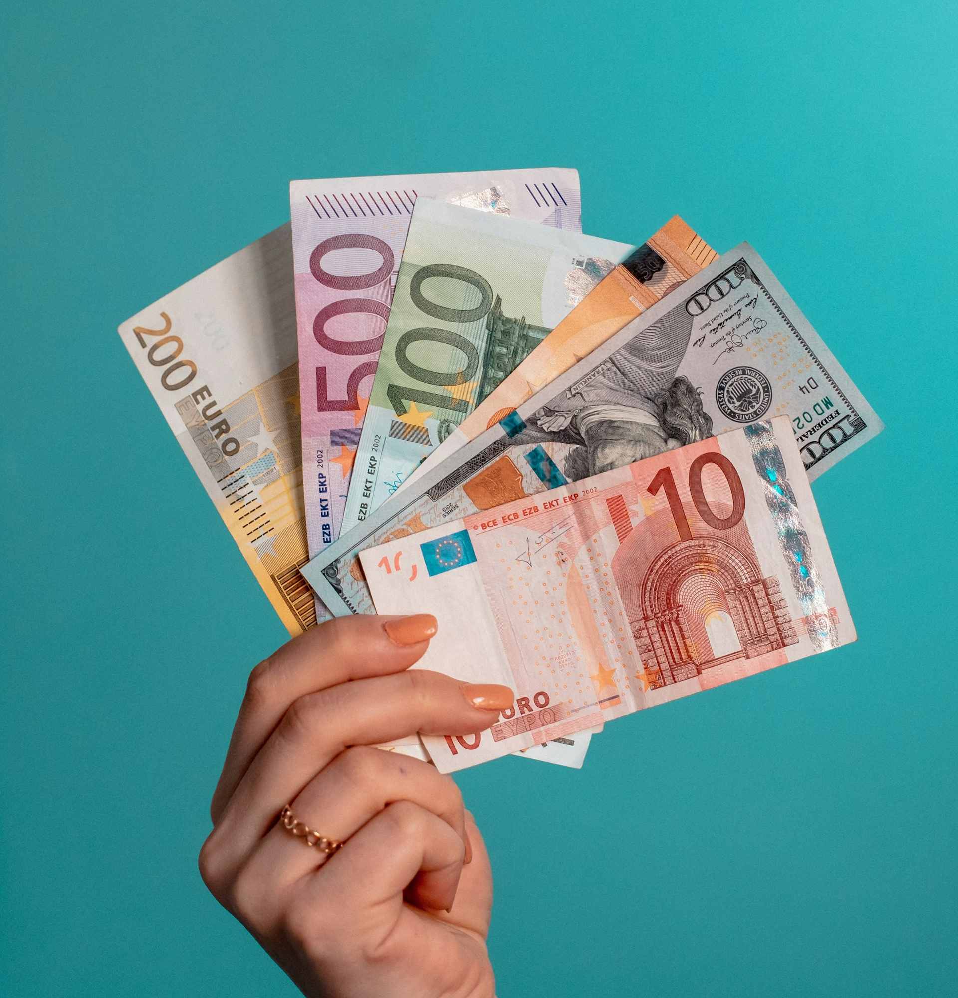 A hand holding a mix of Euro and US dollar banknotes against a turquoise background.