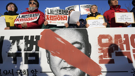 Protesters in South Korea hold banners calling for President Yoon Suk Yeol's resignation, with a large defaced poster of the president in the background.